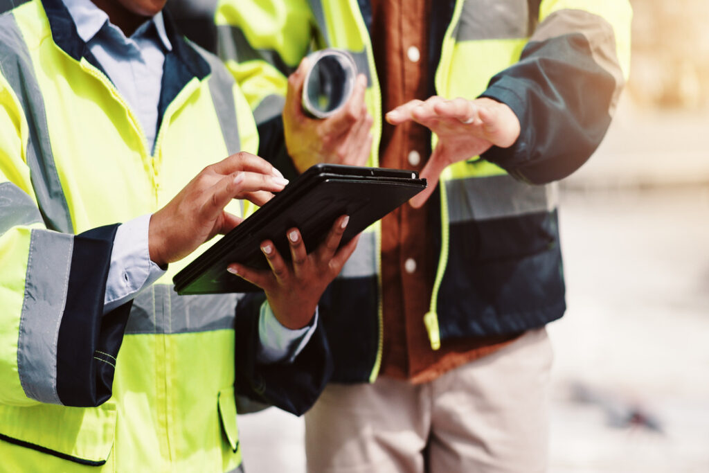 Profissionais em ambiente industrial utilizando coletes de alta visibilidade enquanto realizam inspeção técnica com tablet, representando a avaliação de vestimentas FR e critérios de conformidade em campo.
