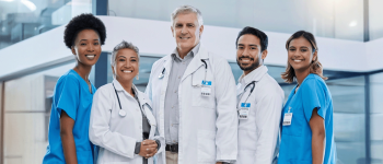 equipe médica composta por duas enfermeiras  vestindo scrubs e 3 medicos de jaleco branco, todos sorriem em frente a um hospital. 