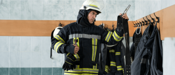homem com vestimenta de proteção tipo de profissionais dos bombeiros próximo à parede onde há varios ganchos com outras vestimentas e equipamentos de segurança.