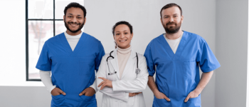 dois homens vestindo scrubbs azuis e uma mulher usando jaleco e um instrumento médico,  todos sorrindo em um corredor cinza claro iluminado.