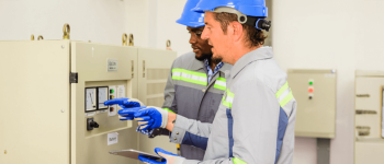 Dois homens uniformizados usando EPI,  capacete, luvas de borracha e uniformes  com faixa refletiva.