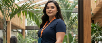 Uma mulher de cabelos escuros sorri para a foto, vestindo uma camiseta azul-marinho profissional com um cordão laranja no pescoço. Ela está em um ambiente moderno de escritório com elementos de madeira e muitas plantas verdes ao fundo.