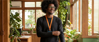 Um homem negro sorridente, usando óculos e blusa preta de manga longa com um crachá de cordão laranja, caminha por um escritório decorado com plantas e acabamentos em madeira.
