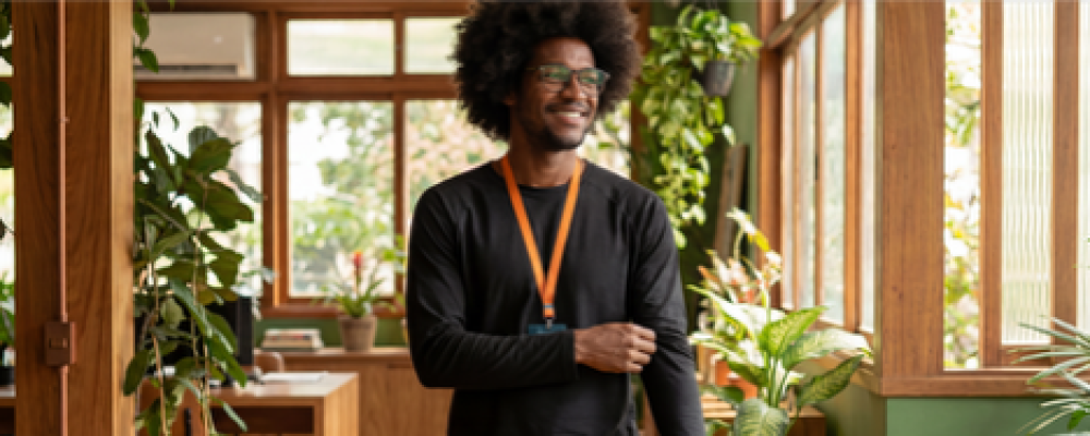 Um homem negro sorridente, usando óculos e blusa preta de manga longa com um crachá de cordão laranja, caminha por um escritório decorado com plantas e acabamentos em madeira.