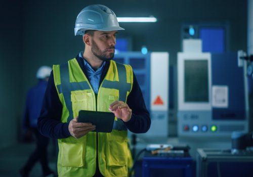 Engenheiro com capacete branco e colete refletivo operando tablet em fábrica